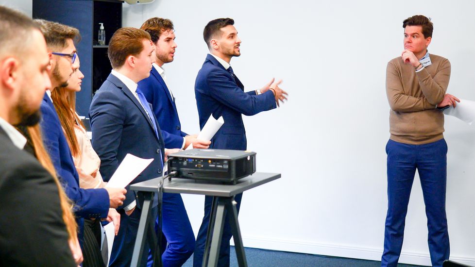 Studierende in einer Veranstaltung Mehrere Studierende stehen nebeneinander. Sie tragen in Business-Kleidung und halten teilweise Notizen in der Hand. Ein Student spricht und zeigt mit der Hand zur Seite. Ein Professor steht mit etwas Abstand seitlich von den Studierenden.