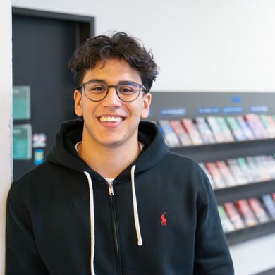 Nicolas Molina Sanchez Auf dem Bild ist Nicolas Molina Sanchez zusehen. Er steht mit der Schulter angelehnt an einer weißen Wand. Im Hintergrund sind bunte Flyer in einem Regal ausgestellt. Nicolas Sanchez trägt eine schwarze Sweatshirt-Jacke und hat braune lockige kurze Haare und eine runde Brille.