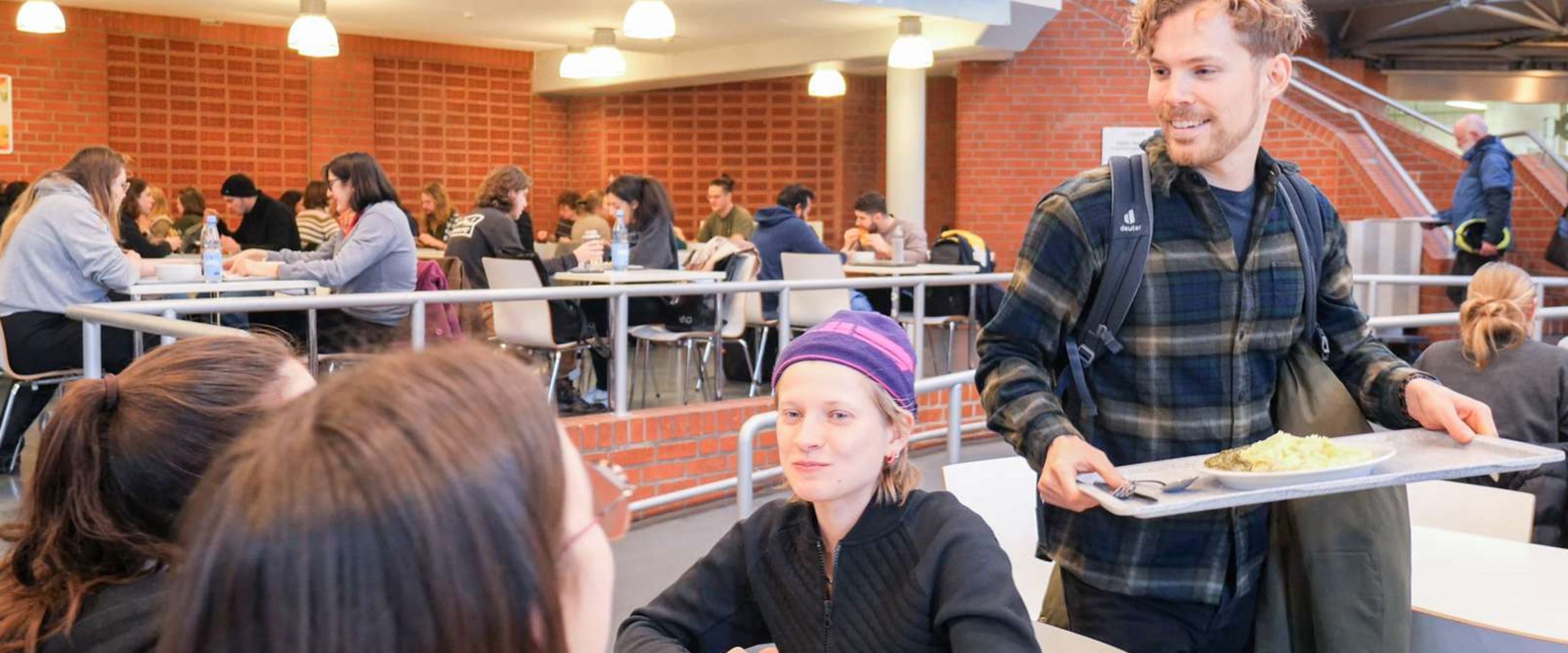 Mensa Essen Auf dem Bils sitzt eine Gruppe Studierenden an einem Tisch in der Mensa. Ein Studierender trägt gerade ein Tablett mit Essen zum Tisch.