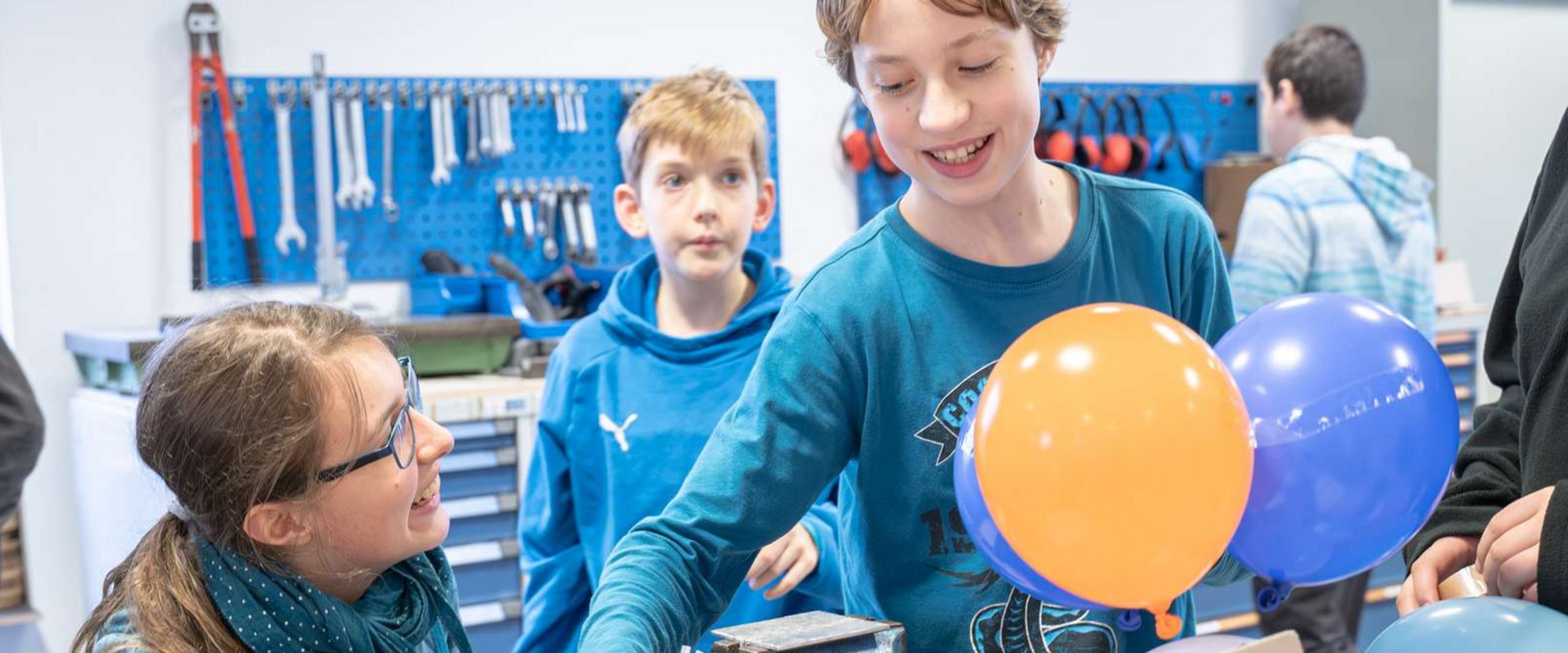 Egg-drop-contest_Skyhub Auf dem Bild sind zwei Schüler und eine Schülerin zu sehen. Gemeinsam basteln sie an einer Konstruktion. Der Junge hält die Konstruktion mit zwei großen Luftballons. Das Mädchen und der andere Junge schauen ihm zu.