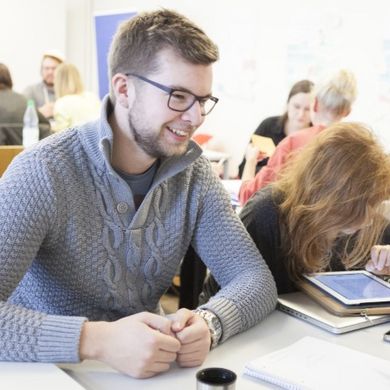 Studierende Drei HSB Studierende sitzen zusammen an einem Tisch. Zwei der drei Studierenden sitzen sich gegenüber und lächeln sich an. Die dritte Studierende blickt vorgebeugt auf ein Tablet. Im Hintergrund sitzen weitere HSB Studierende.