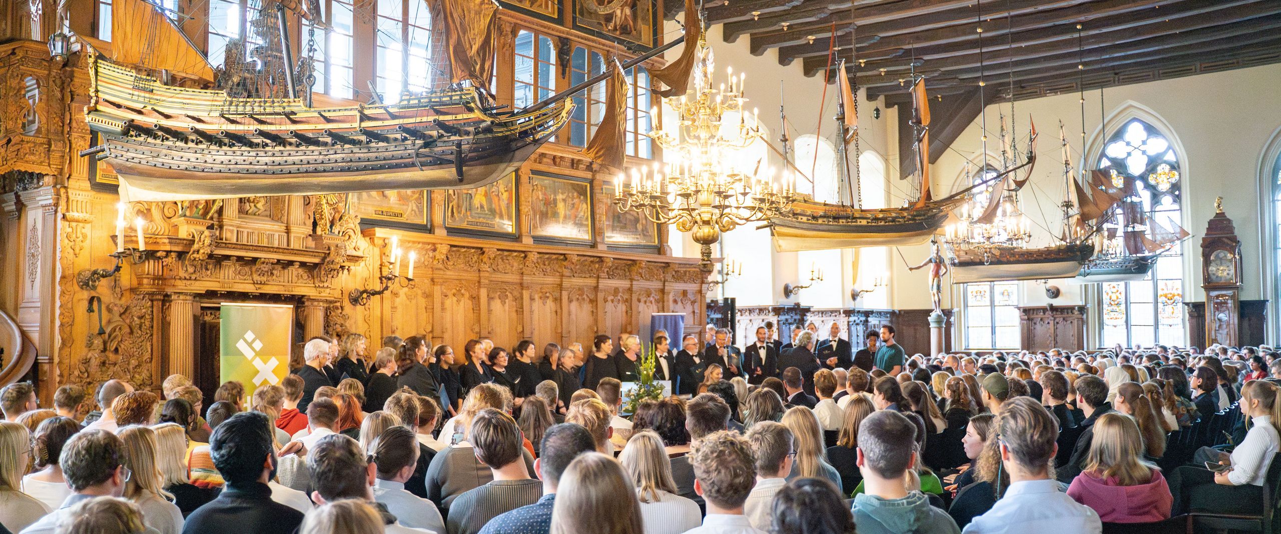 Festassal mit vielen Gästen. Von der Decke hängen alte Holzschiffmodelle. Vorne steht ein Chor.