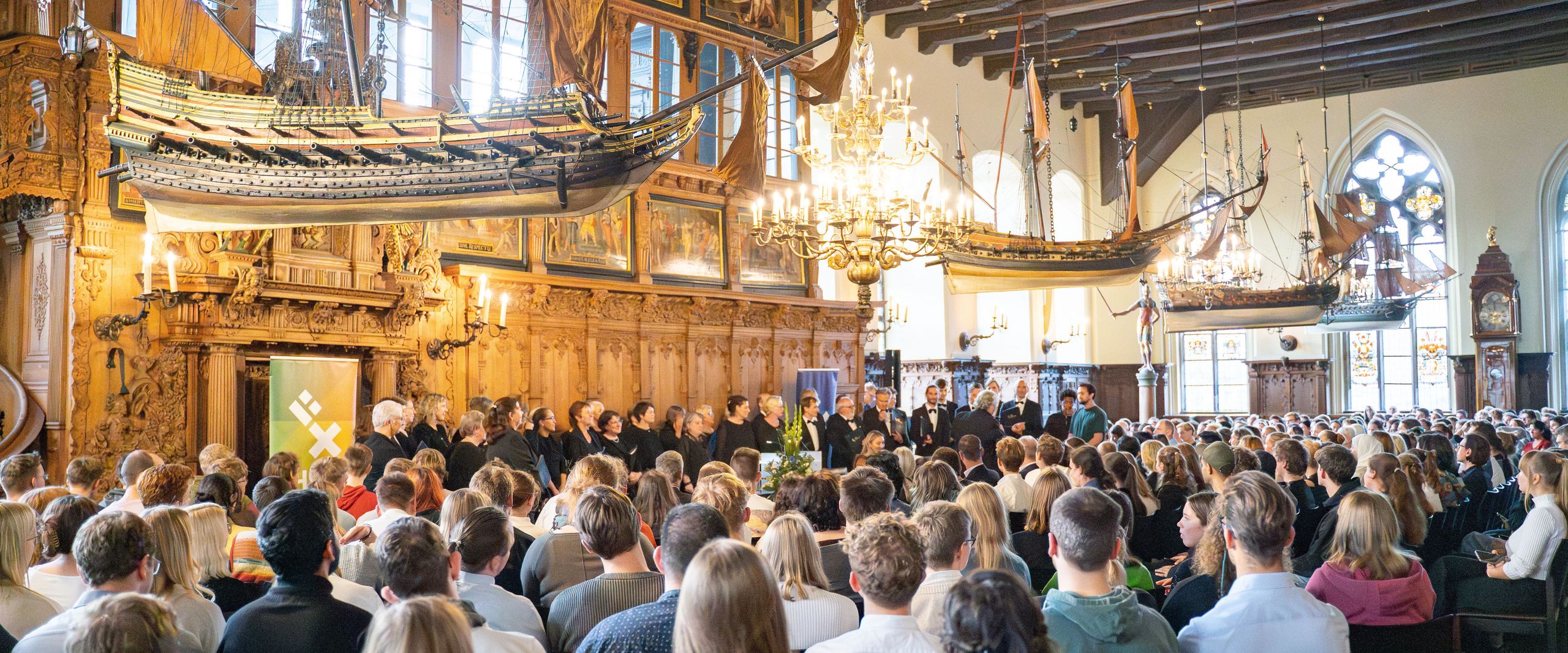 The picture shows the upper town hall, filled with HSB students and staff. Model ships hang from the ceiling. A choir stands in front 