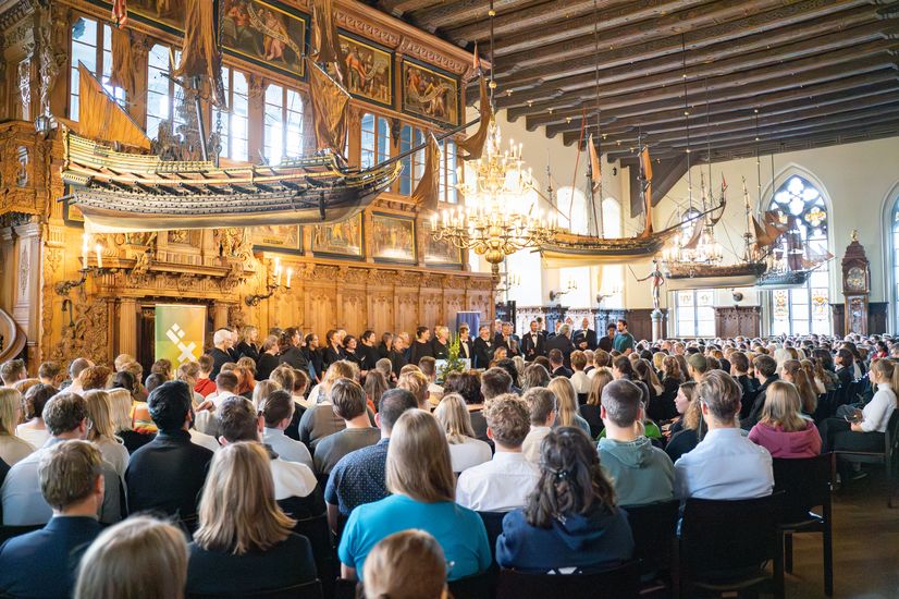 Festassal mit vielen Gästen. Von der Decke hängen alte Holzschiffmodelle. Vorne steht ein Chor.