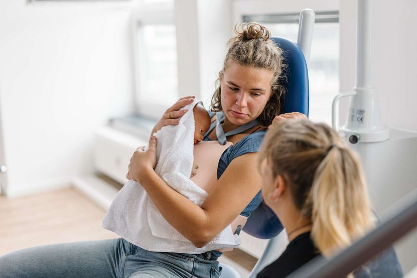 Studiengang Hebammen Auf dem Bild ist eine Studentin des Studiengangs Hebammen zu sehen, die eine Situation kurz nach der Geburt simuliert. Sie sitzt auf einem Stuhl und hält eine Puppe im Arm, die ein Neugeborenes simuliert.