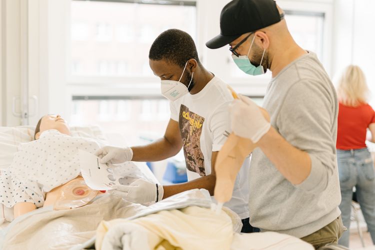 Pflegestudierende im Simulationszentrum des Studiengangs. Pflegestudierende üben an einem Krankenhausbett mit einer Puppe im Simulationszentrum des Studiengangs.