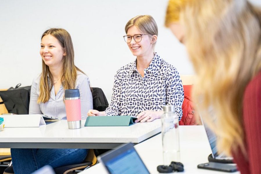 Studentinnen im Seminar Zwei Studentinnen sitzen nebeneinander an einem Tisch in einem Seminarraum. Sie lachen und schauen nach vorn. Im Vordergrund des Fotos sieht man unscharf eine weitere Studentin von der Seite.