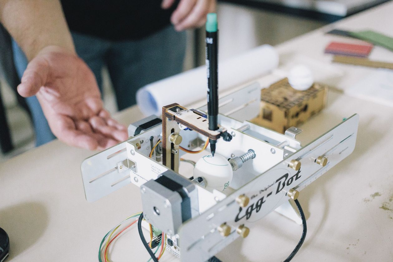 Praxis in der Vorlesung Eine Hand deutet auf einen Roboter, der Eier bemalen kann.